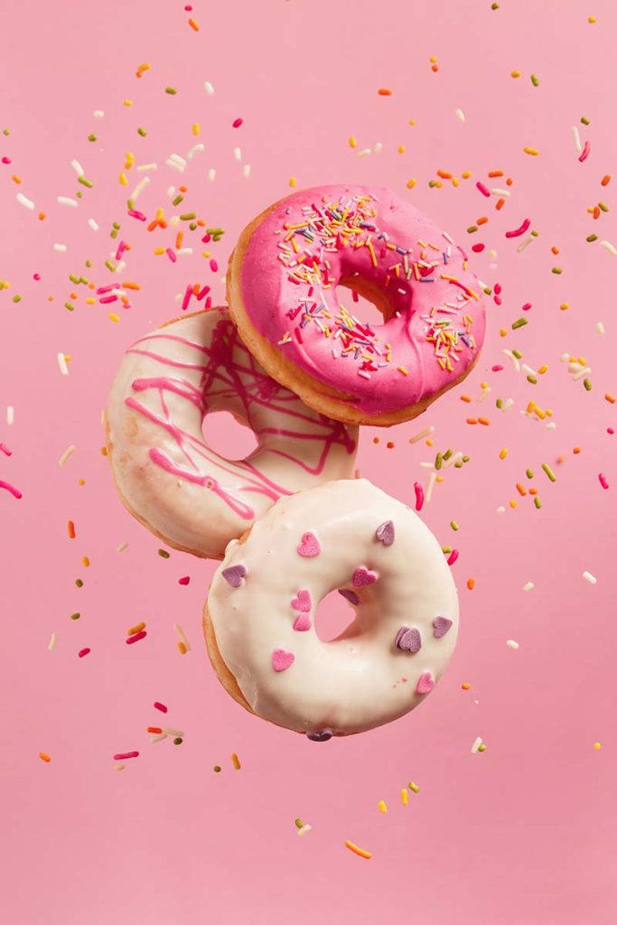 Stylised photo of three donuts falling past the camera, with a pink background. There are sprinkles falling down behind the donuts.